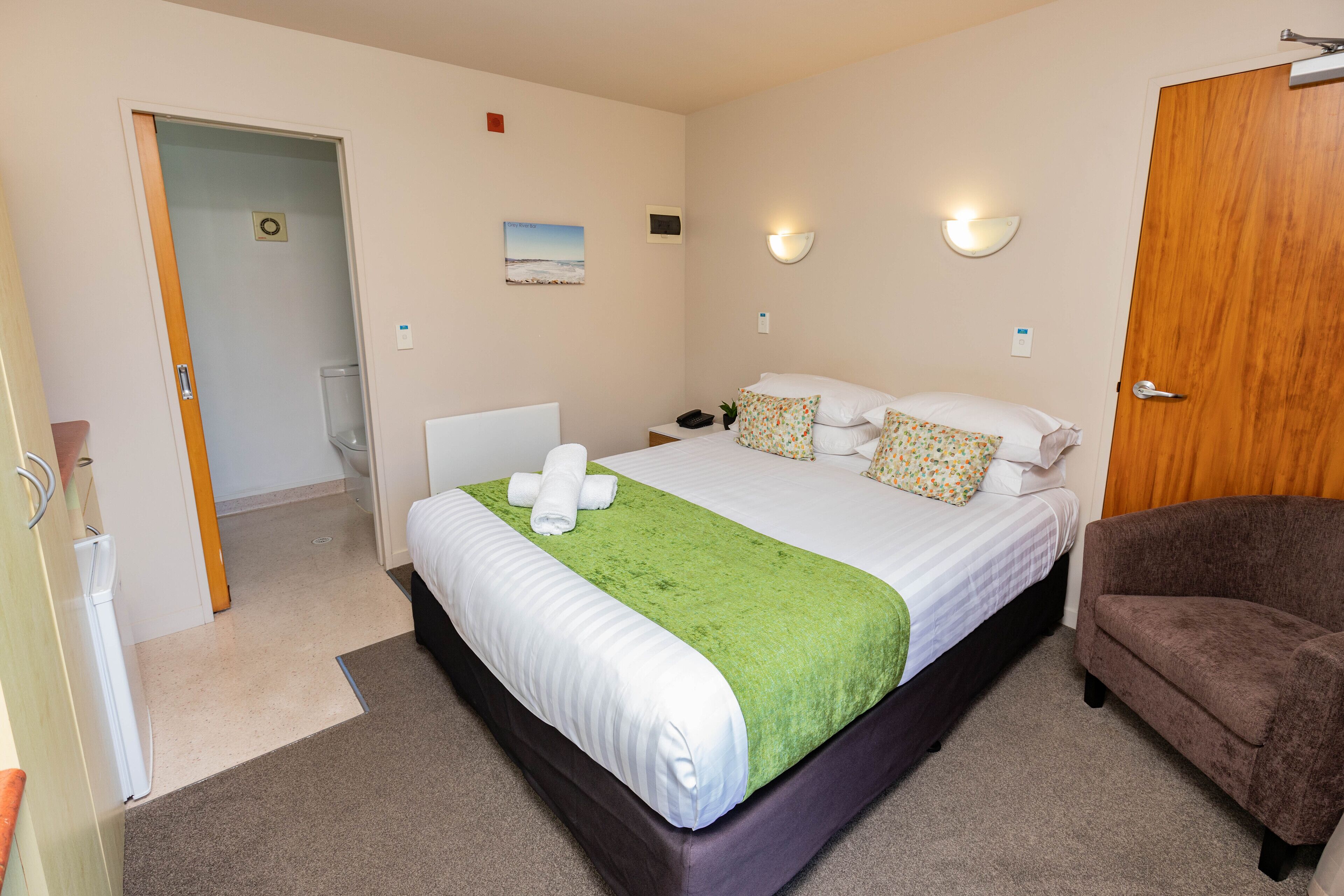 A hotel room with a large bed, a brown armchair, a bedside table with a lamp, and a wall-mounted light fixture.
