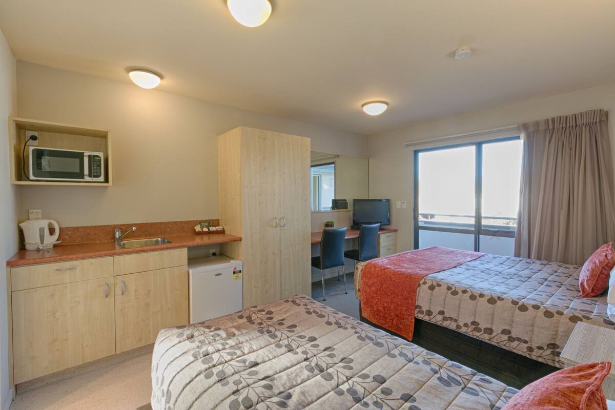 A hotel room with two beds, a kitchenette, a desk, a TV, and a large window with curtains.
