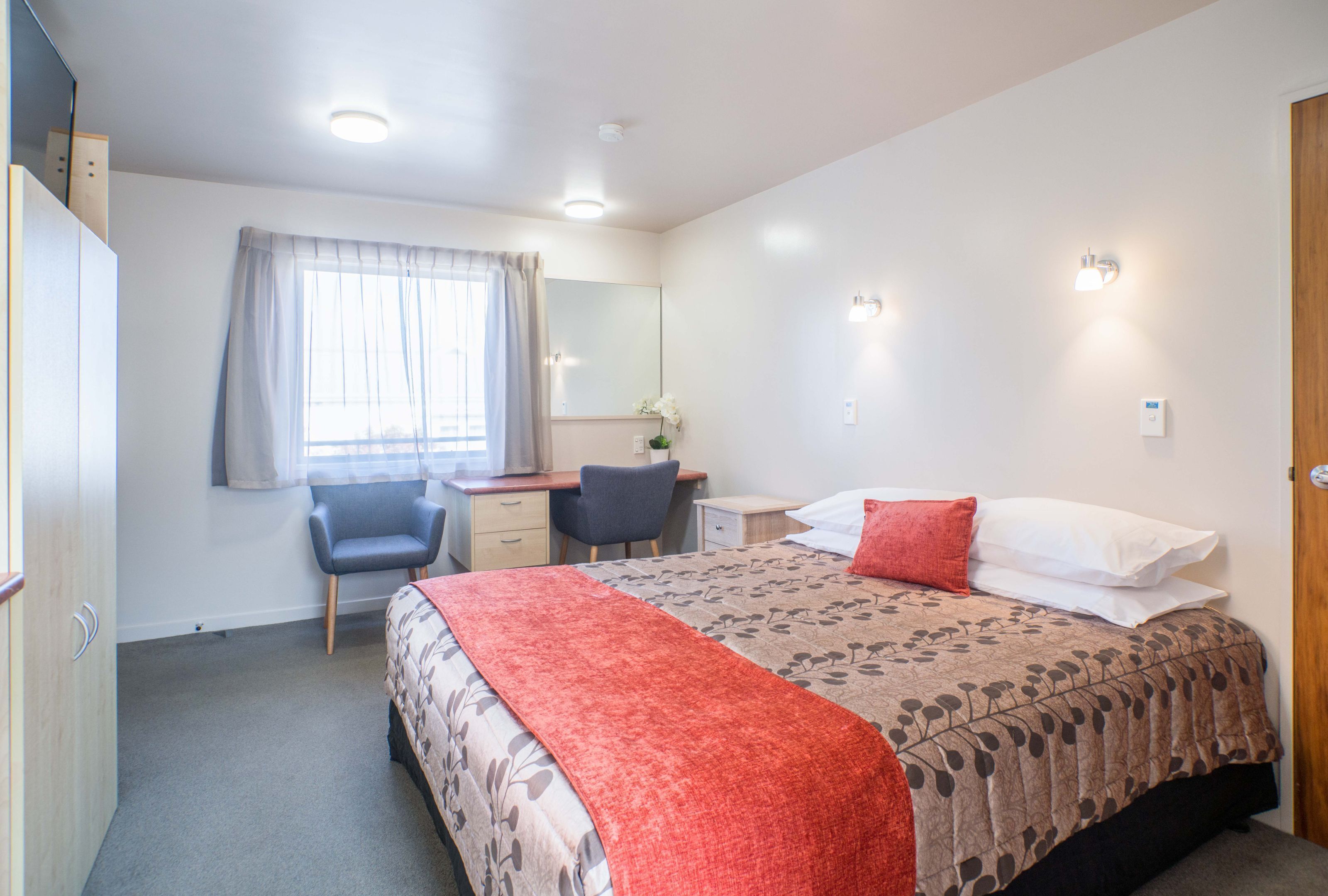 Quarto de hotel com cama, secretária, cadeira, guarda-roupa e janela com cortinas.