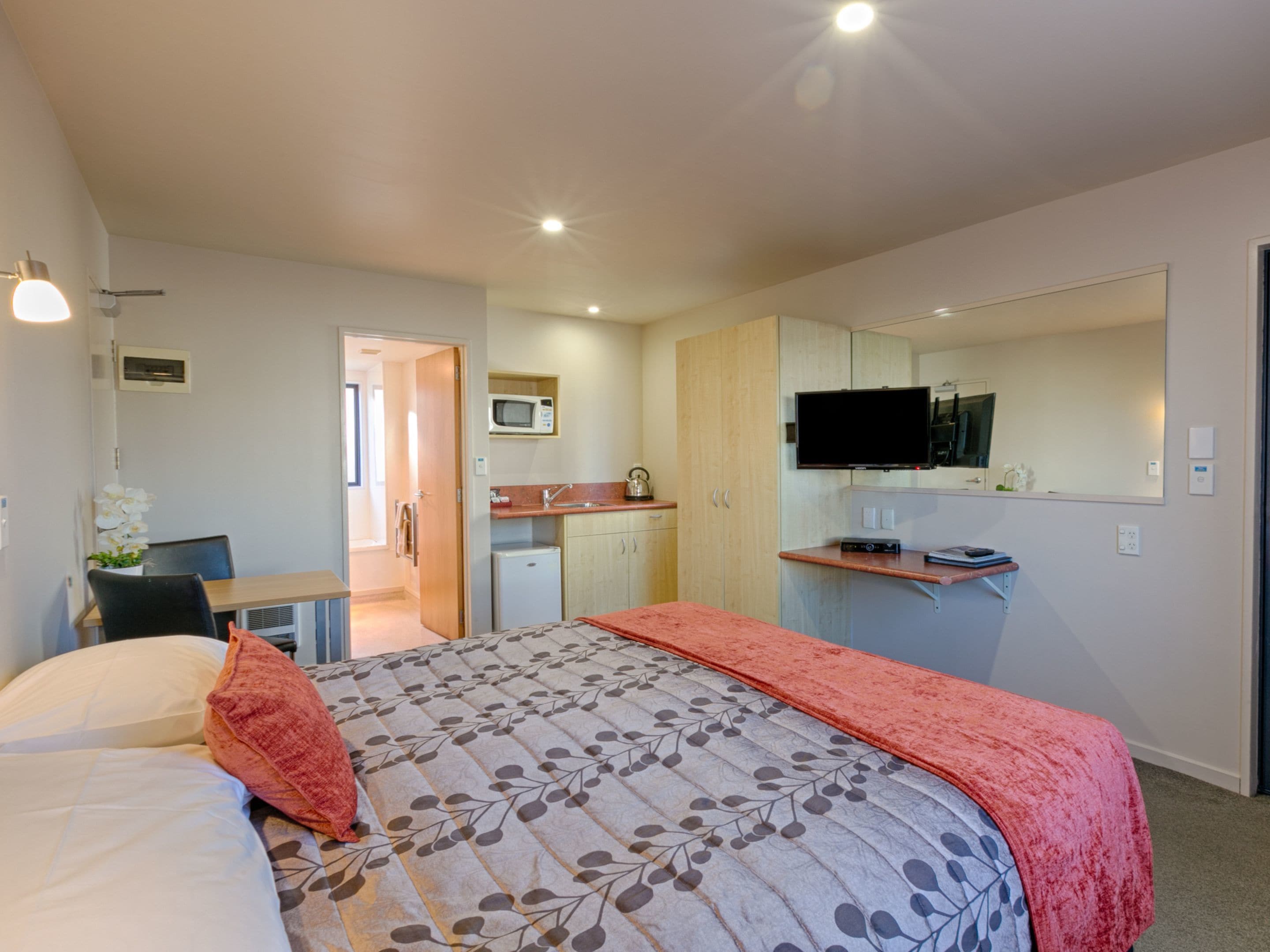A hotel room with a bed, a desk, a kitchenette, and a TV.