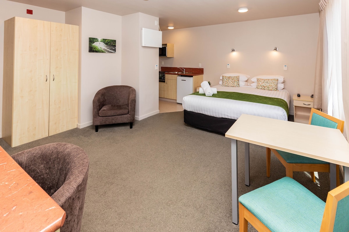 A compact hotel room with a bed, a small kitchenette, a seating area with a table and chairs, and a wardrobe.