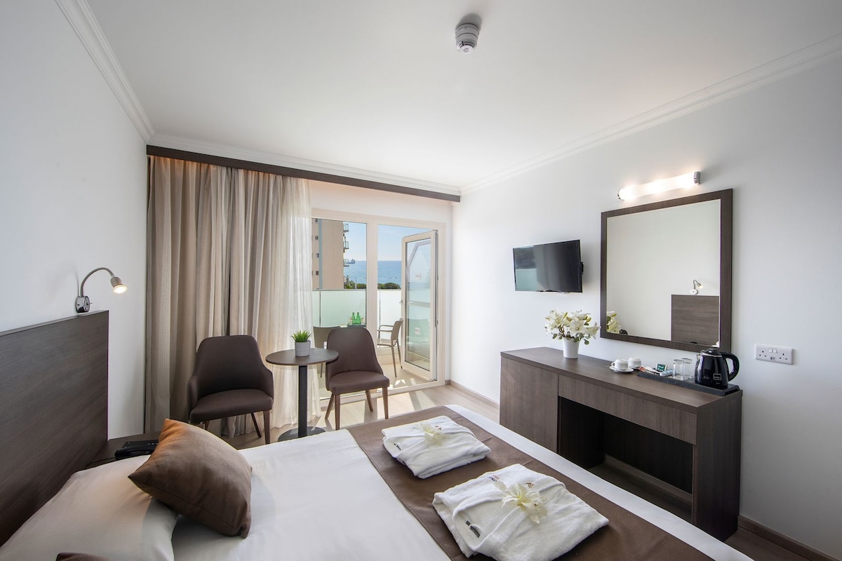 A hotel room with a bed, a desk, a chair, a TV, a mirror, a balcony with a view, and a coffee maker.