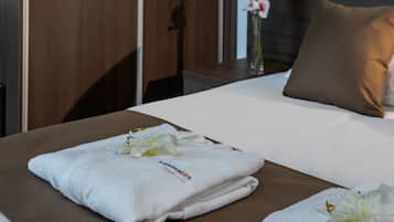 A neatly made bed with a folded towel and a flower, a brown pillow, and a vase with flowers in the background.