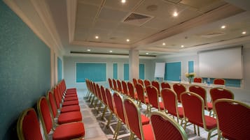A conference room with rows of red chairs and a long table, equipped with a projector screen and a water dispenser.