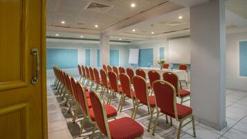 Una sala conferenze con file di sedie rosse, un tavolo bianco con un vaso di fiori e una lavagna.