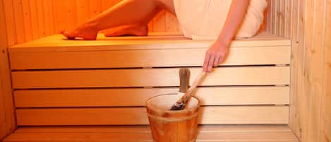 Una sauna de madera con una persona sentada en un banco, un cubo de madera y un mazo de madera.