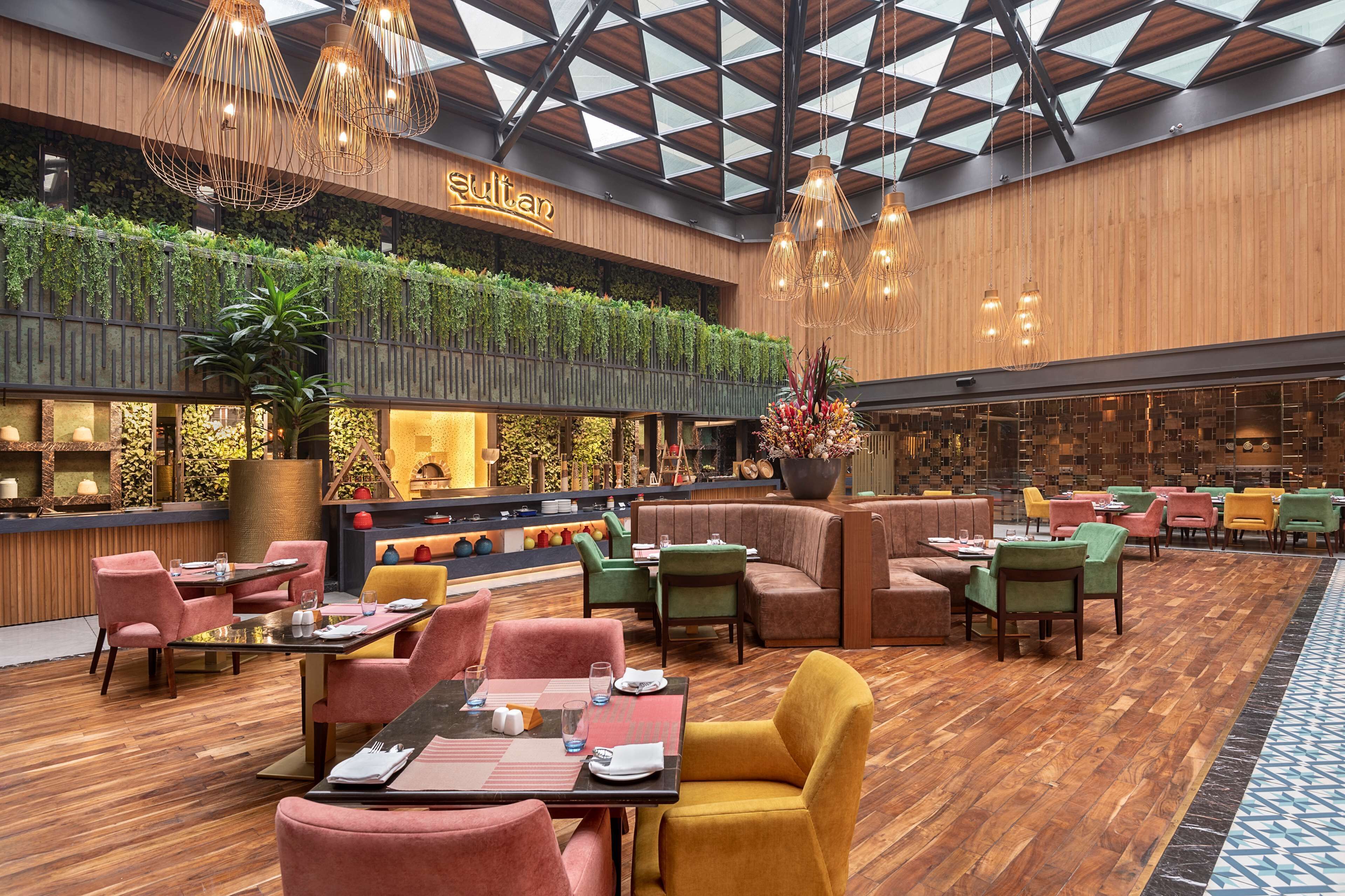 A spacious indoor dining area with multiple seating options, a high ceiling with a geometric pattern, and a green wall with plants.