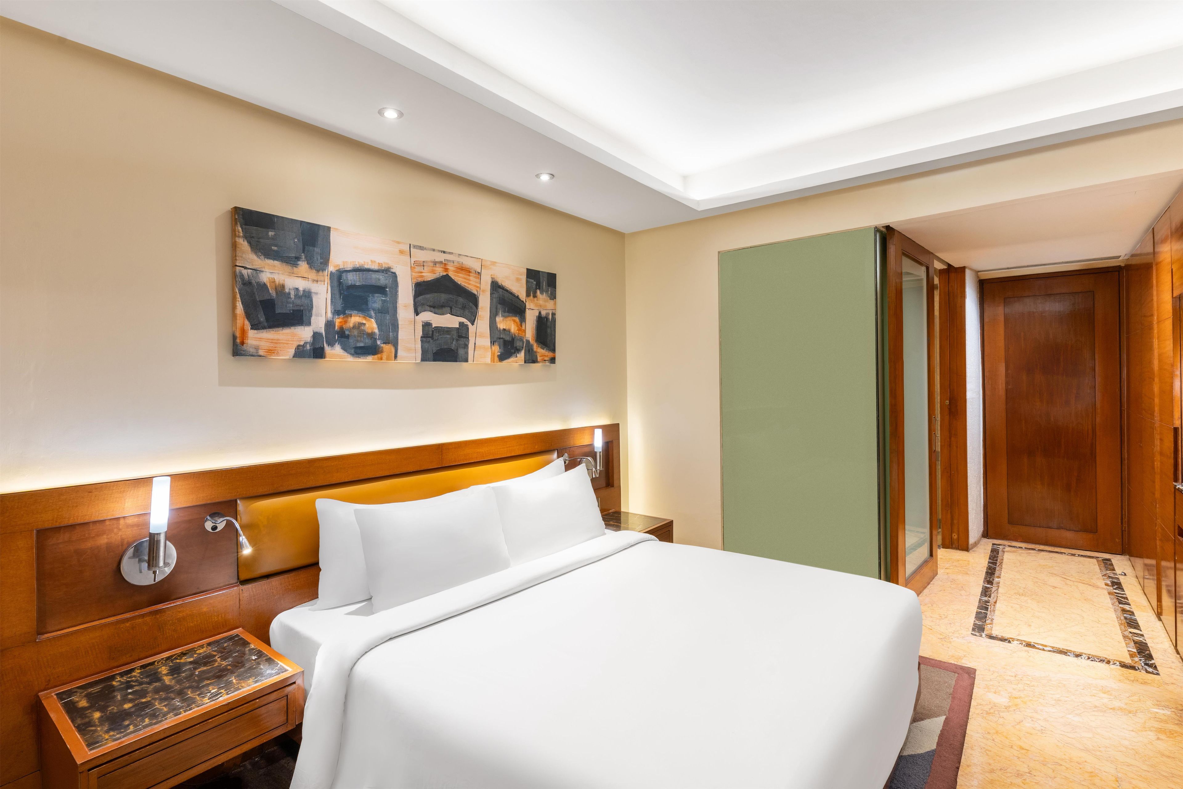A hotel room with a large bed, a wooden headboard, a nightstand, a bedside table, and a wall-mounted artwork.