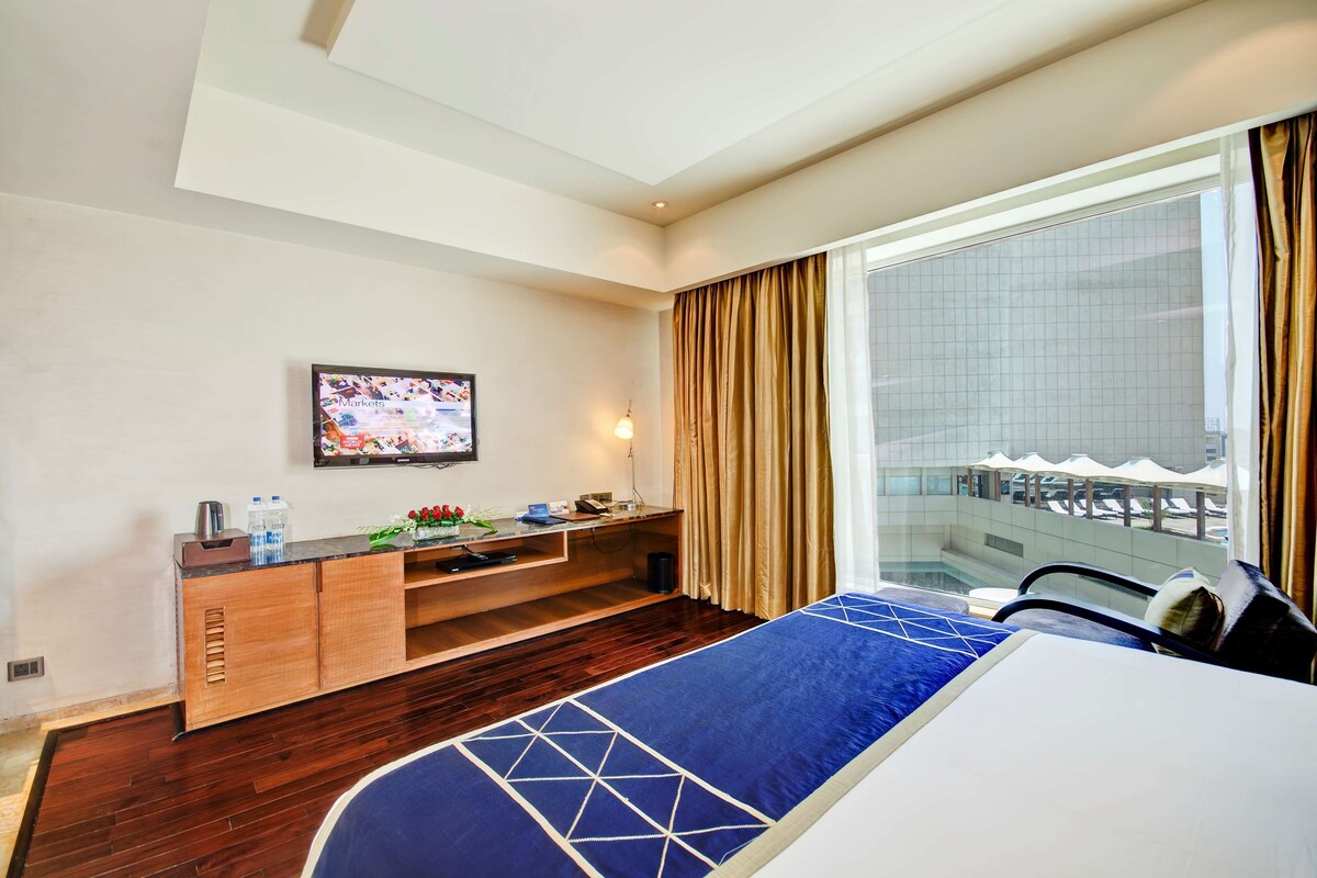Hotelkamer met een bed, bureau met televisie, raam met gordijnen en uitzicht op een zwembad en gebouw.