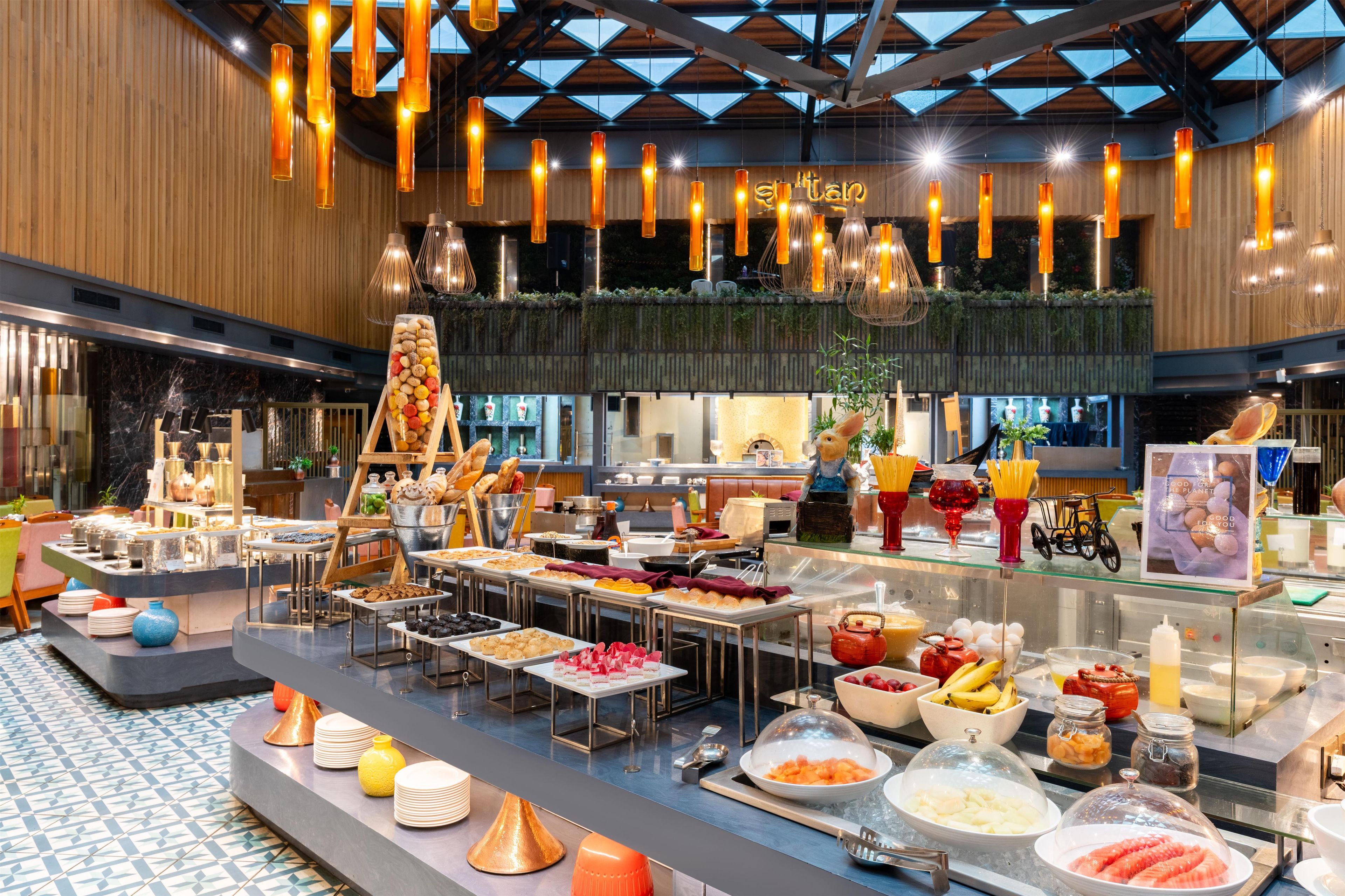 A buffet area with various food items and a salad bar, under a modern ceiling with hanging lights.