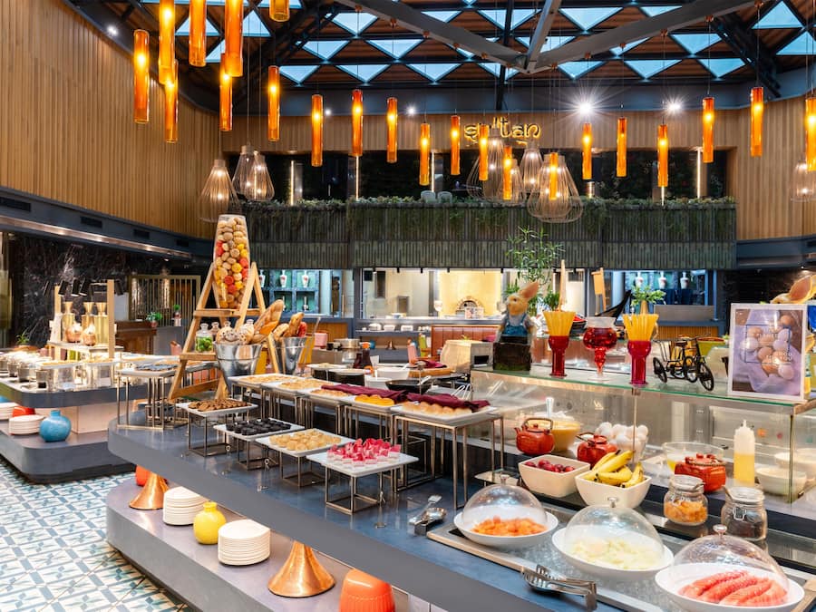 A buffet area with various food items and a salad bar, under a modern ceiling with hanging lights.