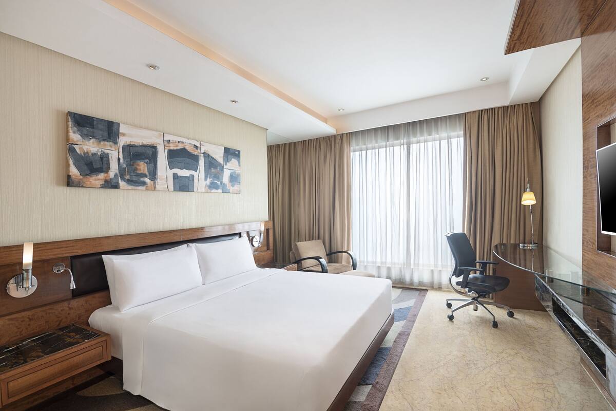 Hotelkamer met een groot bed, een bureau, een flatscreen televisie, een stoel en een groot raam met gordijnen.
