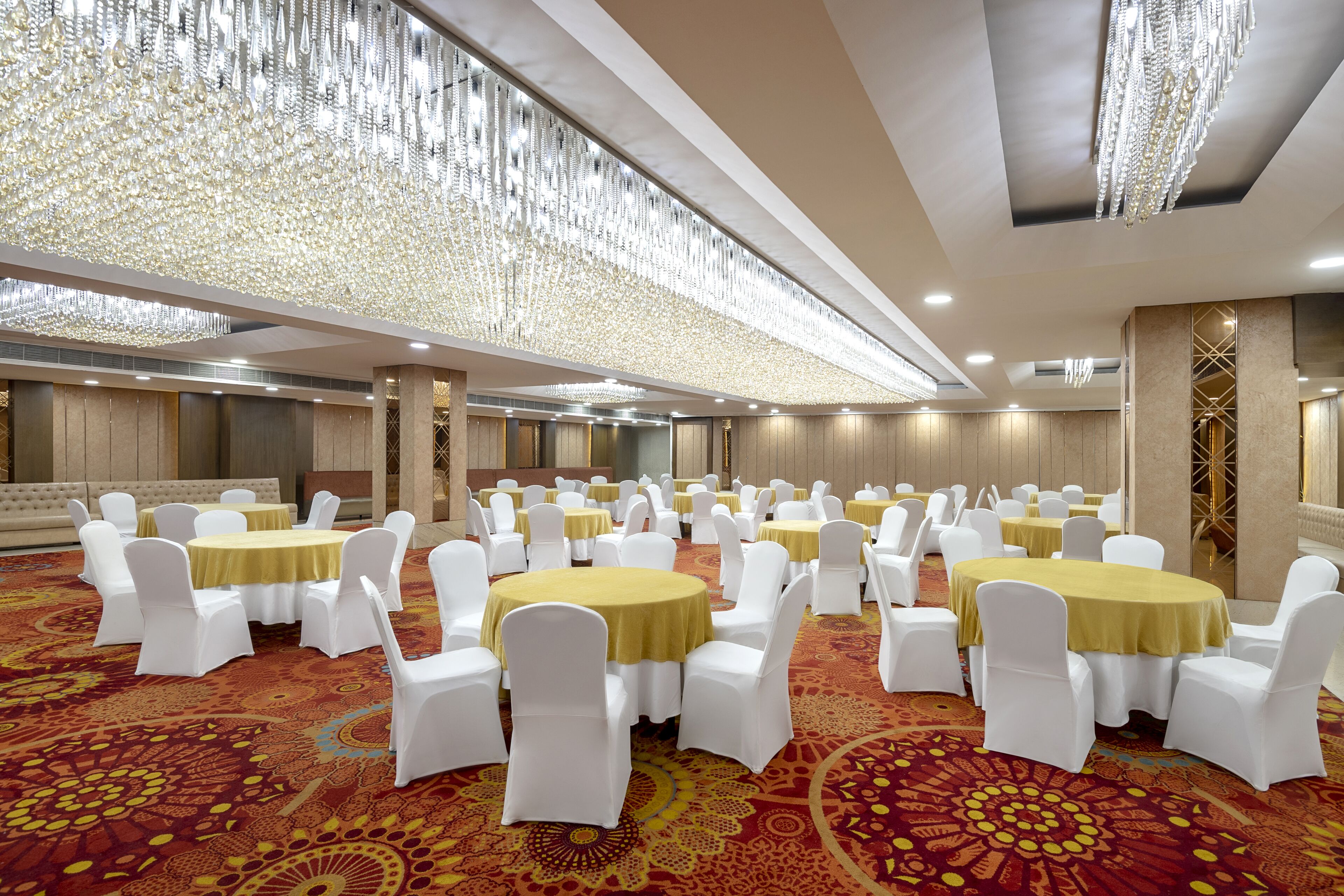 A banquet hall with round tables covered with yellow tablecloths, surrounded by white chairs, and a patterned carpet.