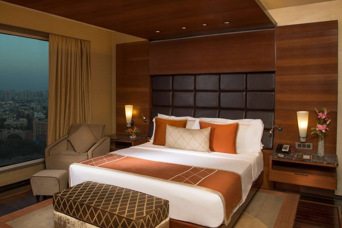 Een moderne hotelkamer met een groot bed, uitzicht over de stad, een zithoek en een nachtkastje met een lamp.