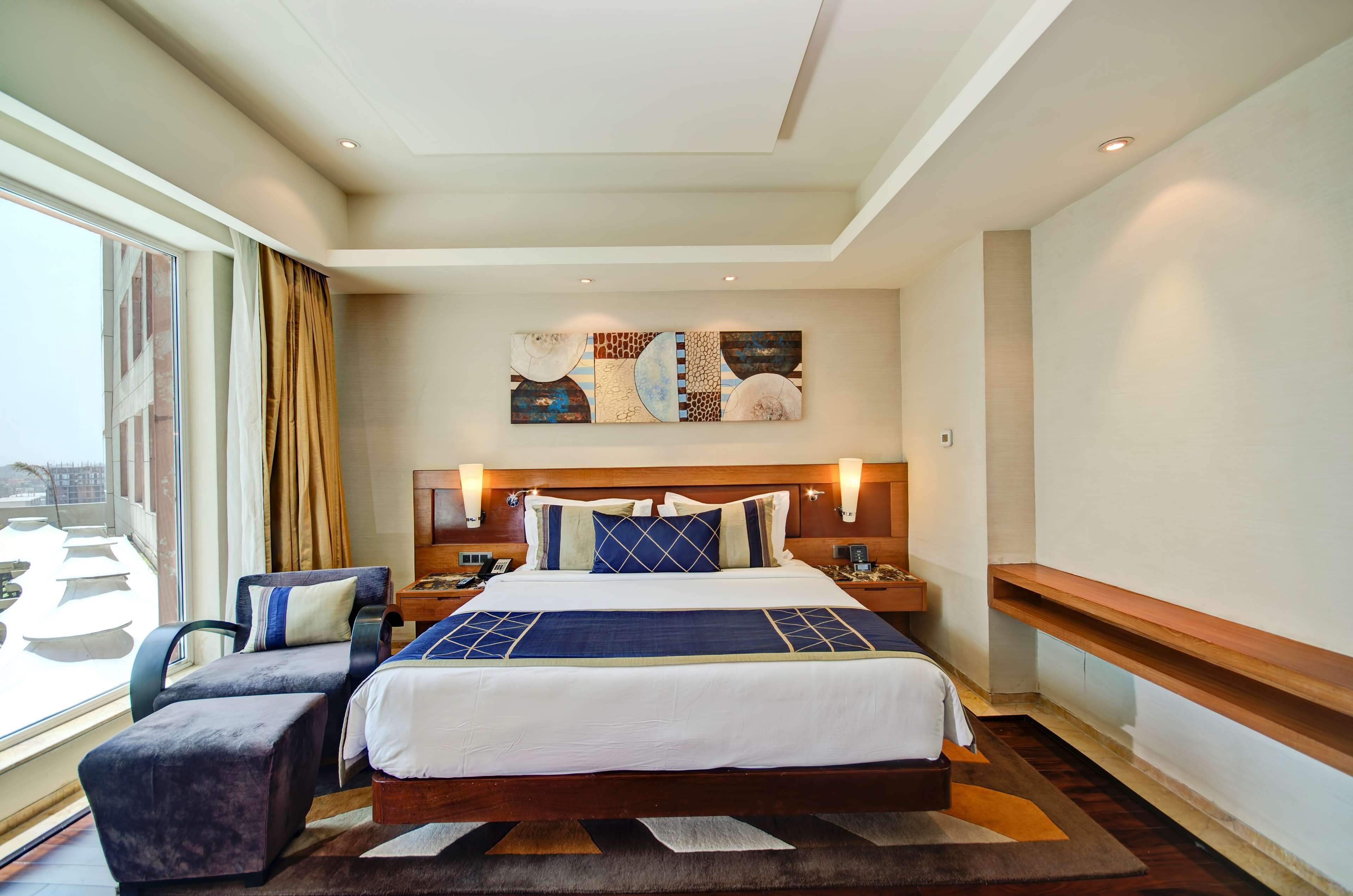Ein Hotelzimmer mit einem großen Bett, einem Schreibtisch, einem Stuhl, einem fenster mit Vorhängen und Blick auf die Stadt.
