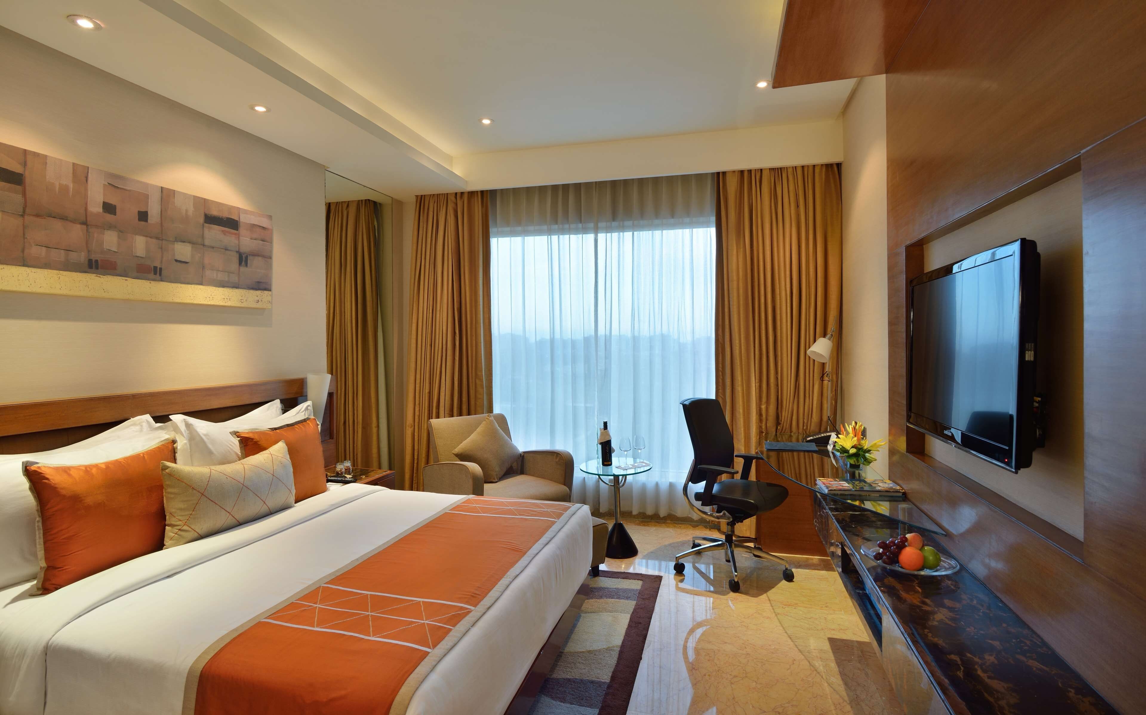 A hotel room with a large bed, a desk with a TV, a chair, and a view of the city through the window.