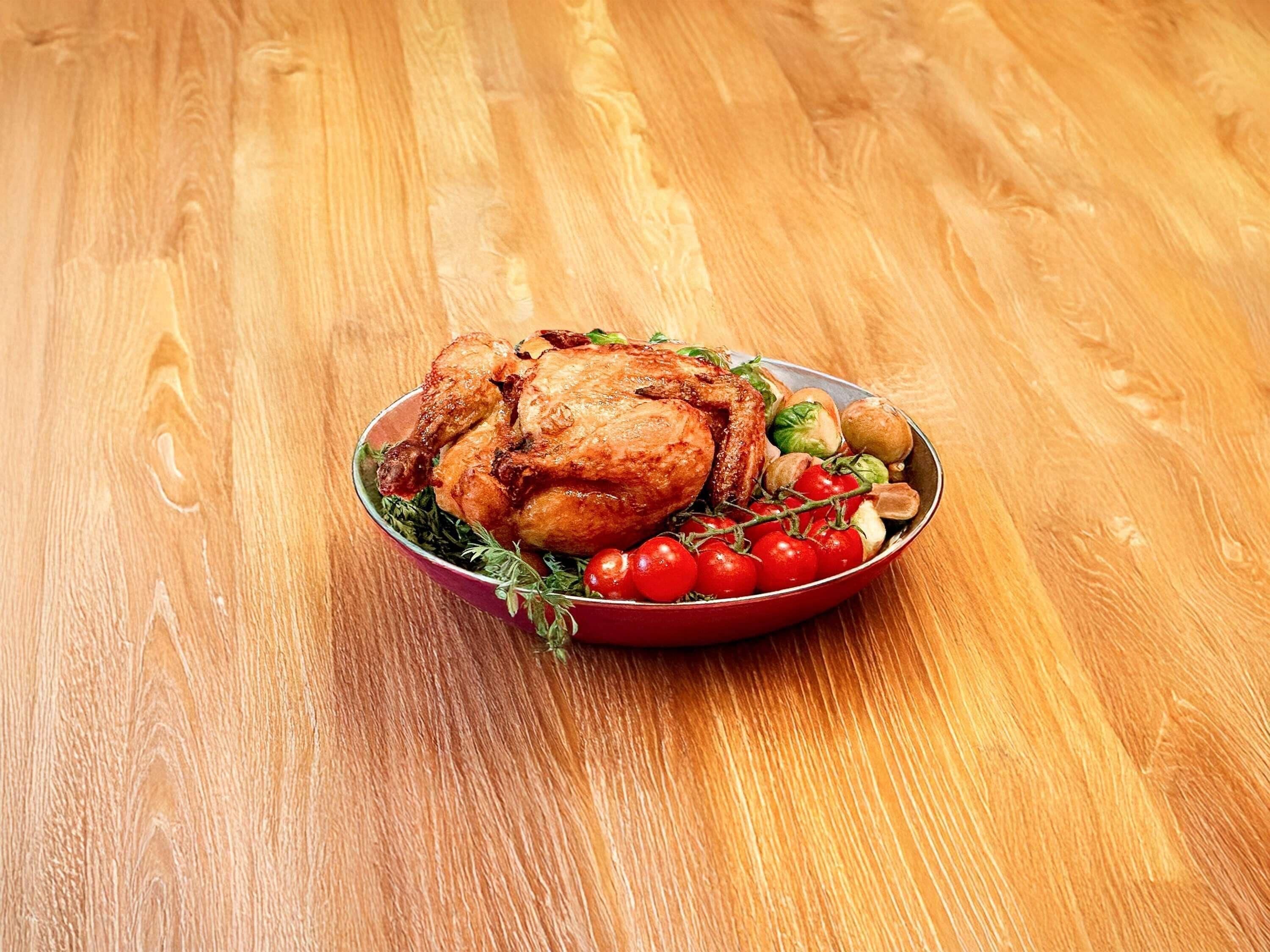 A roasted chicken on a plate with vegetables and herbs, placed on a wooden table.