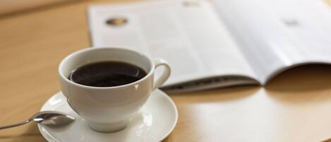A white cup of coffee on a saucer with a spoon, placed on a wooden table next to an open book.