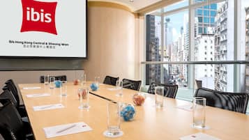 A conference room with a large screen displaying the ibis logo, a long table with chairs, and a city view through large windows.