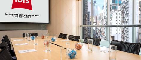 A conference room with a large screen displaying the ibis logo, a long table with chairs, and a city view through large windows.