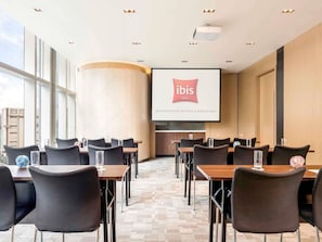 A conference room with a large screen displaying the ibis Hotels logo, multiple tables with chairs, and a view of the city through large windows. - ibis Hong Kong Central And Sheung Wan (Hong Kong)