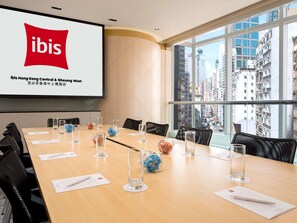 Una sala de conferencias con una pantalla grande mostrando el logotipo de ibis, una mesa larga con sillas y vistas a la ciudad a través de amplios ventanales.