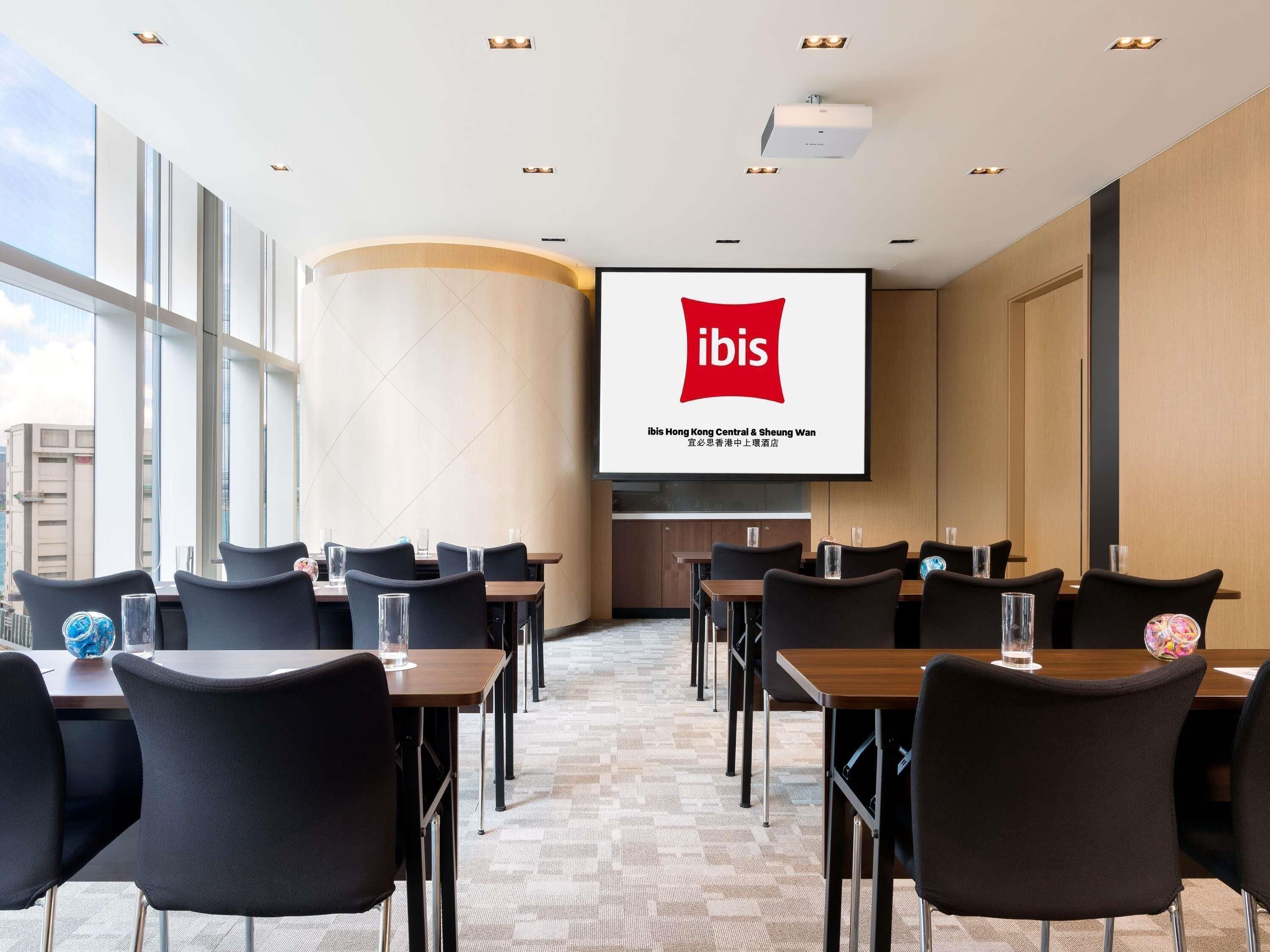 A conference room with a large screen displaying the ibis logo, multiple tables with chairs, and a view of the city through large windows.