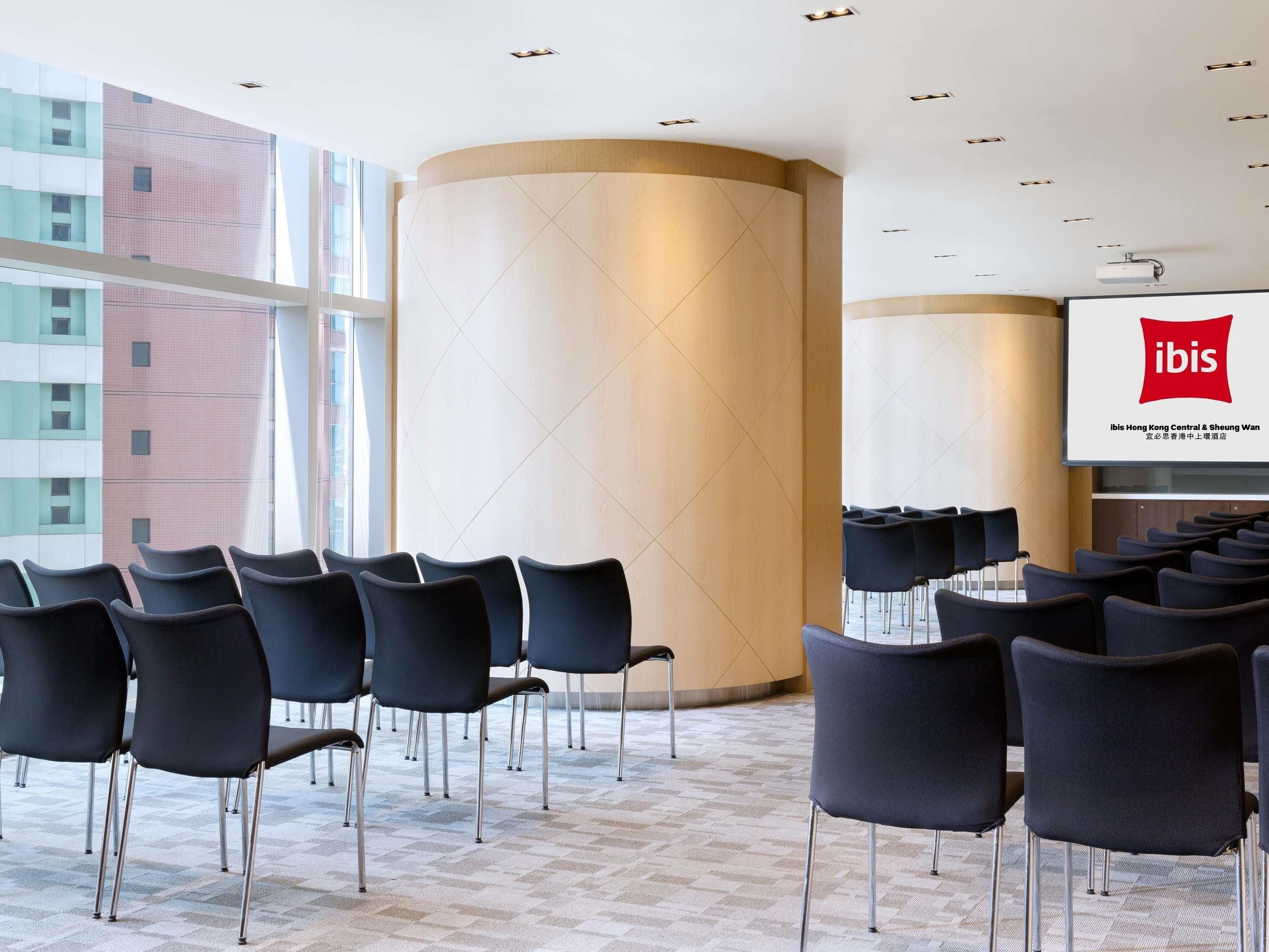A conference room with rows of chairs, a large screen displaying the ibis logo, and a view of a building outside.