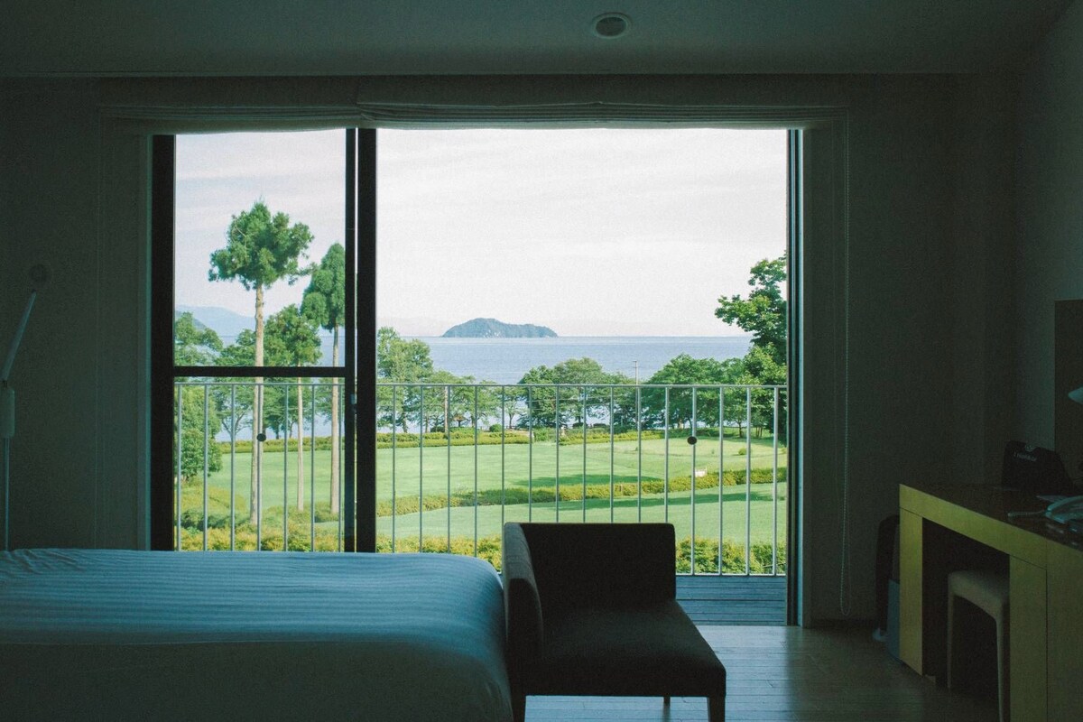 A bedroom with a large window, a bed, a chair, and a desk.