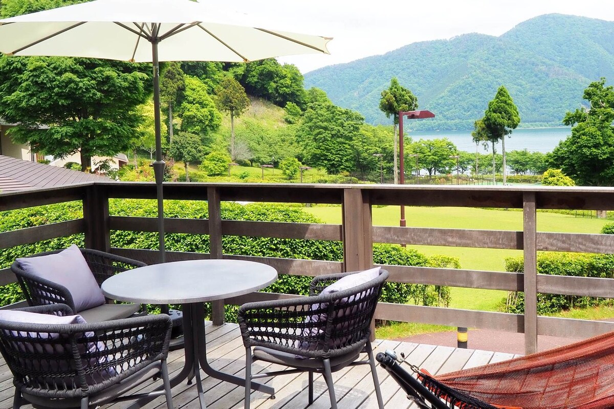 A wooden deck with a table and chairs, a hammock, and a view of a lake and mountains.