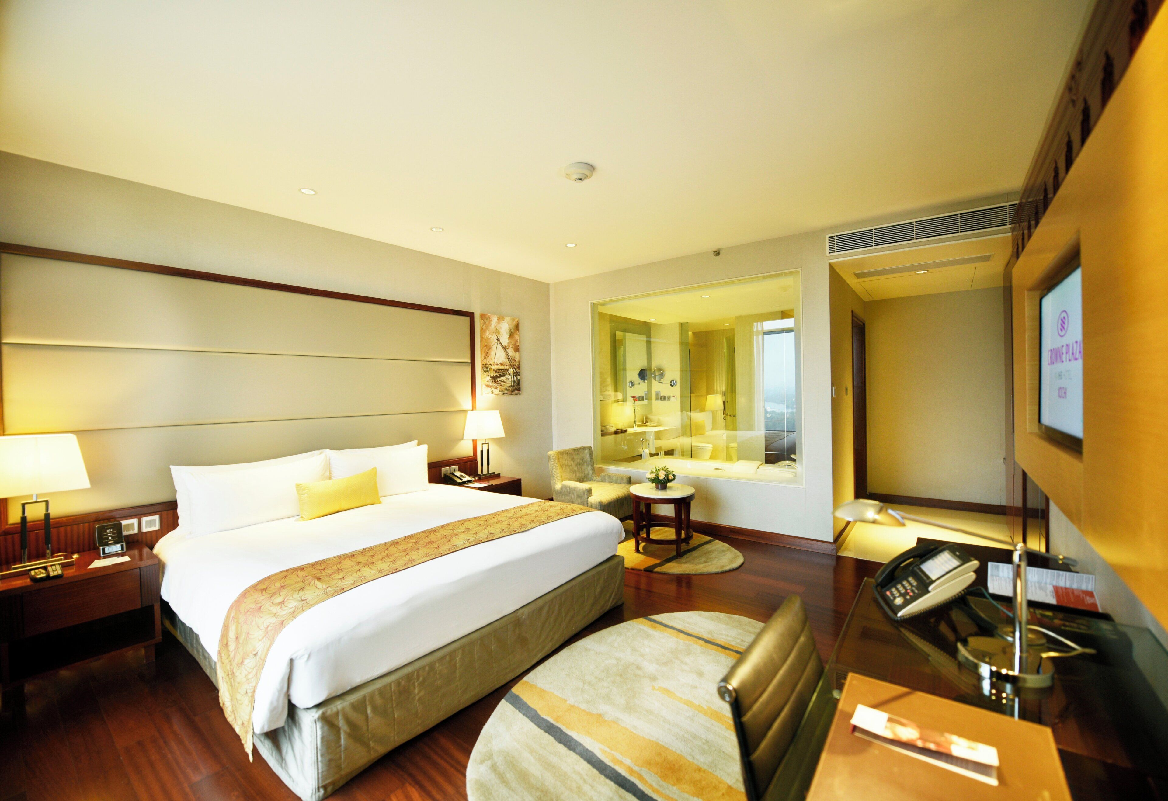 A hotel room with a large bed, a sitting area, a bathroom with a mirror, and a television.