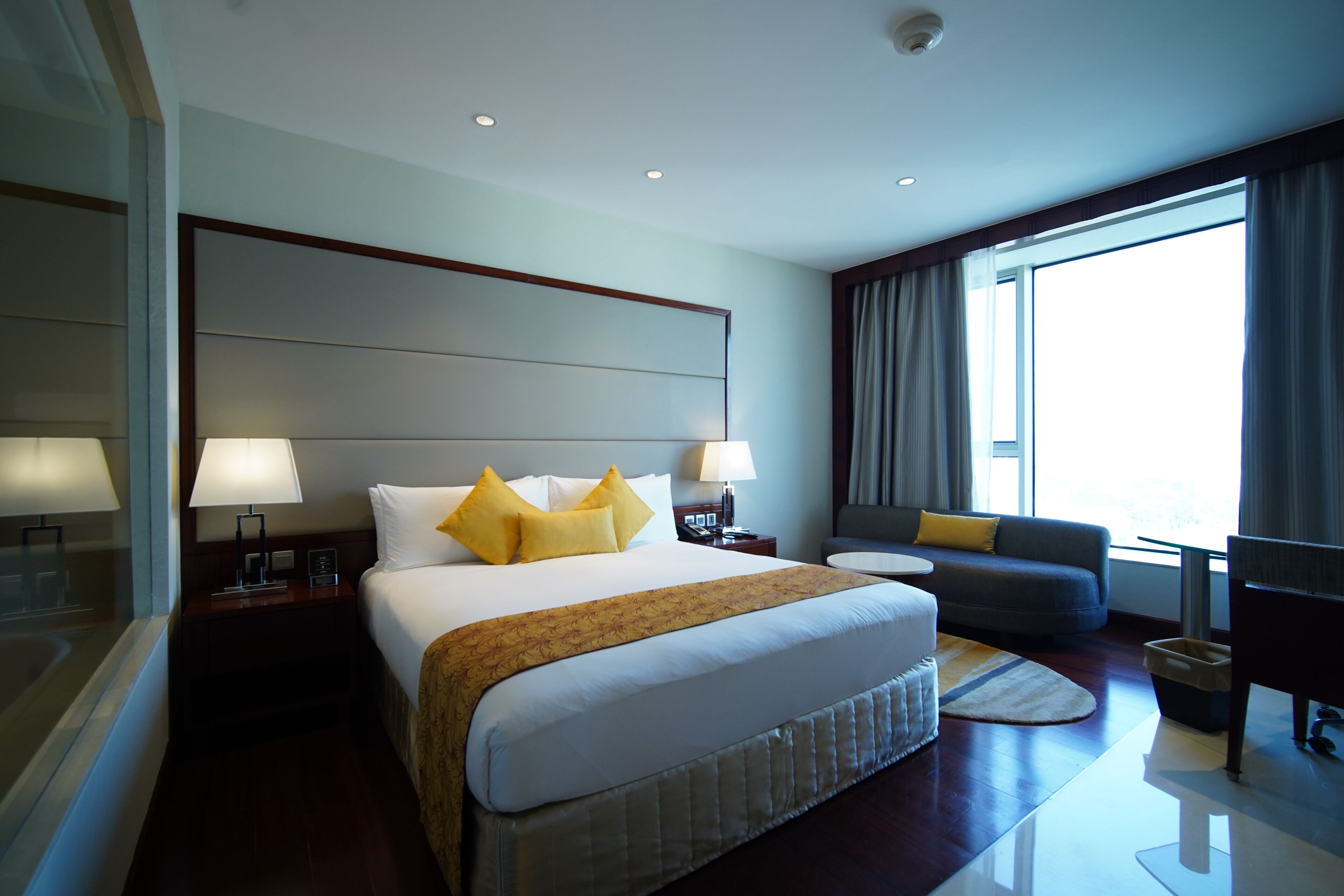 Una camera d'hotel con un letto grande, due comodini con lampade, un divano, un piccolo tavolino rotondo e un'ampia finestra con tende.