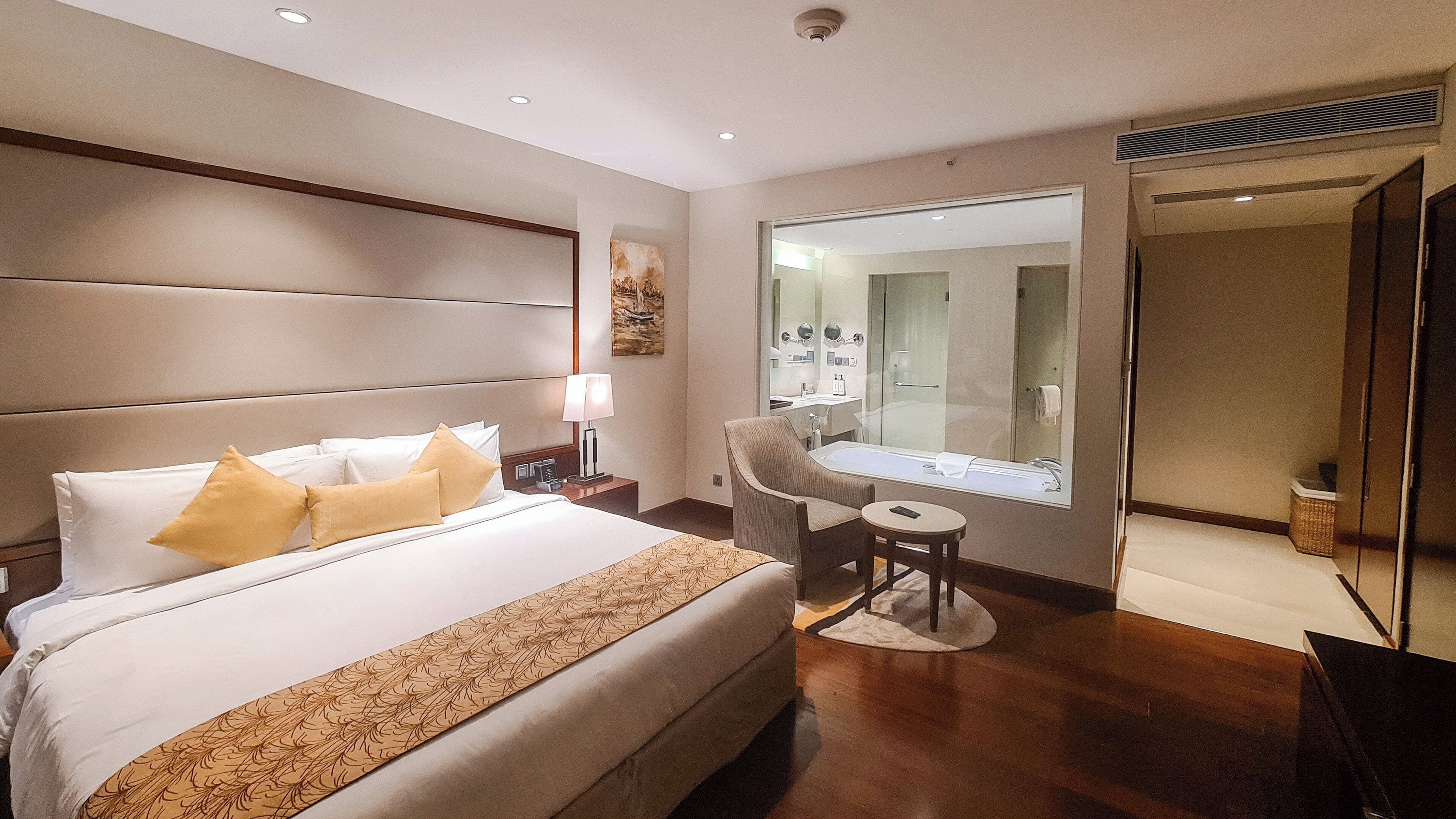 Een hotelkamer met een groot bed, een zithoek met een stoel en een klein tafeltje, een grote spiegel en een badkamer zichtbaar door een glazen deur.