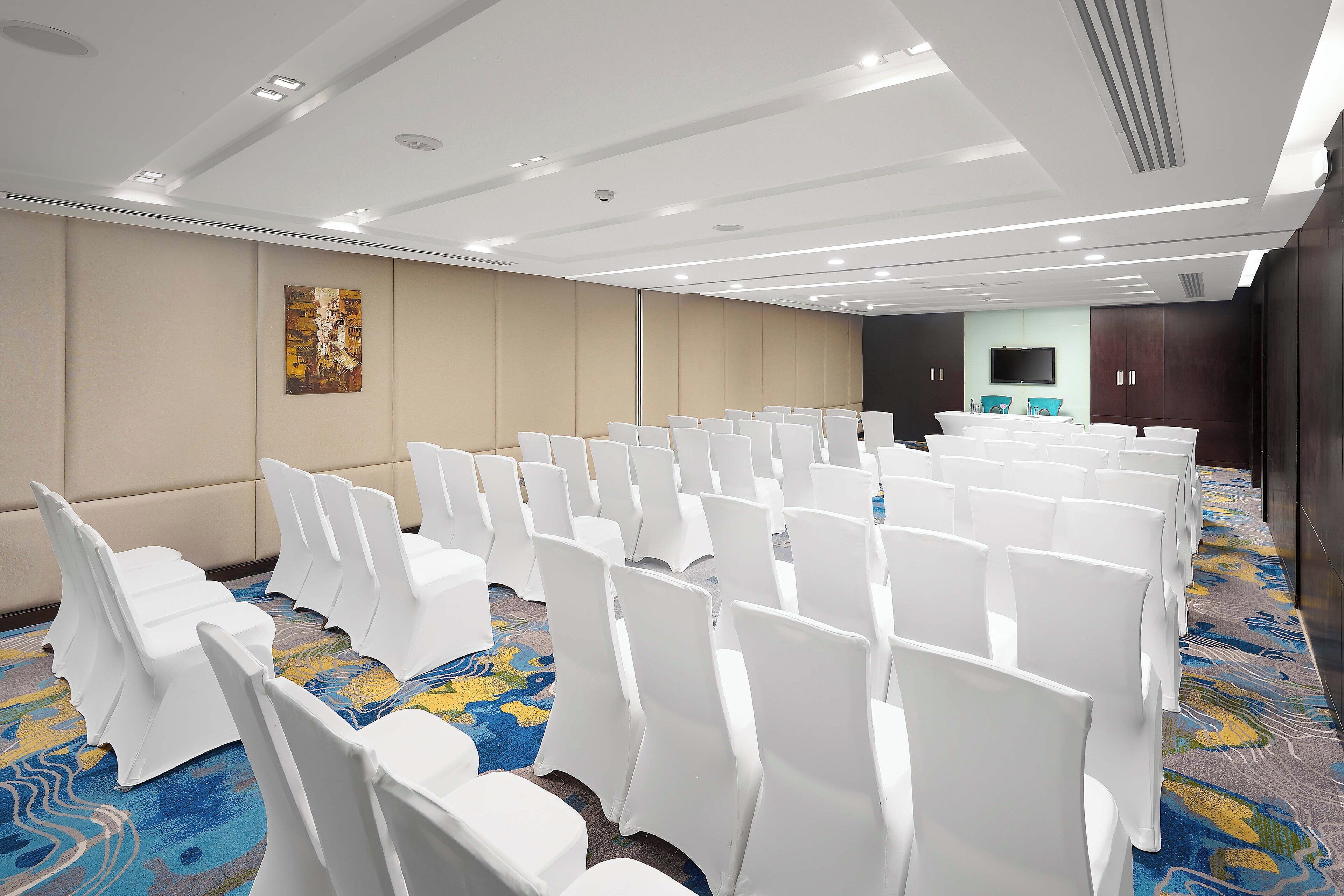 A conference room with rows of white chairs, a carpeted floor, and a flat-screen TV on the wall.