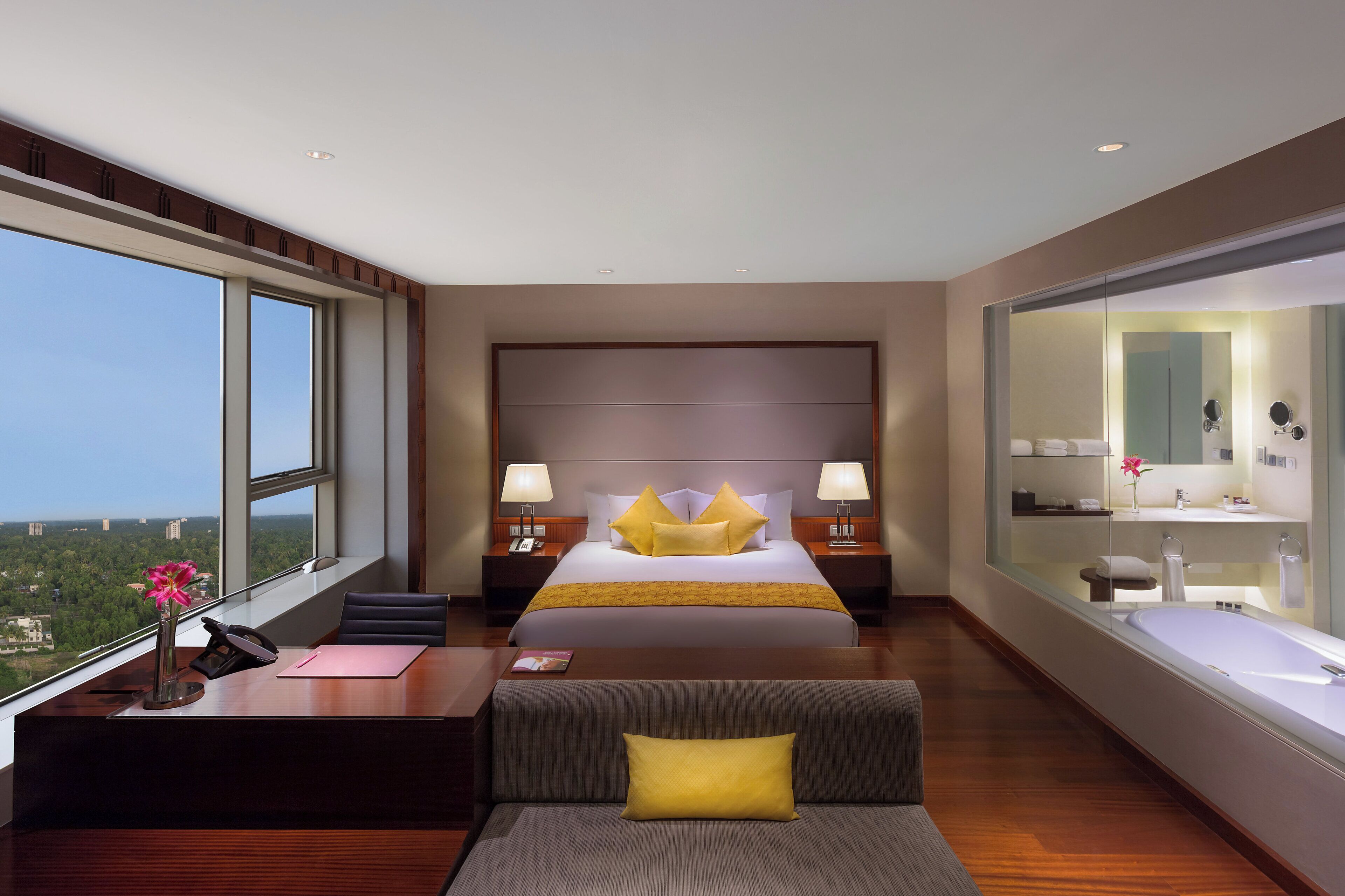 A modern hotel room with a large bed, a desk, a sofa, a view of the city, and a bathroom with a large mirror.