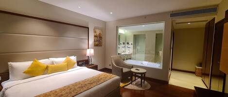 A hotel room with a large bed, a chair, a small table, a mirror, and a bathroom visible through the glass door.