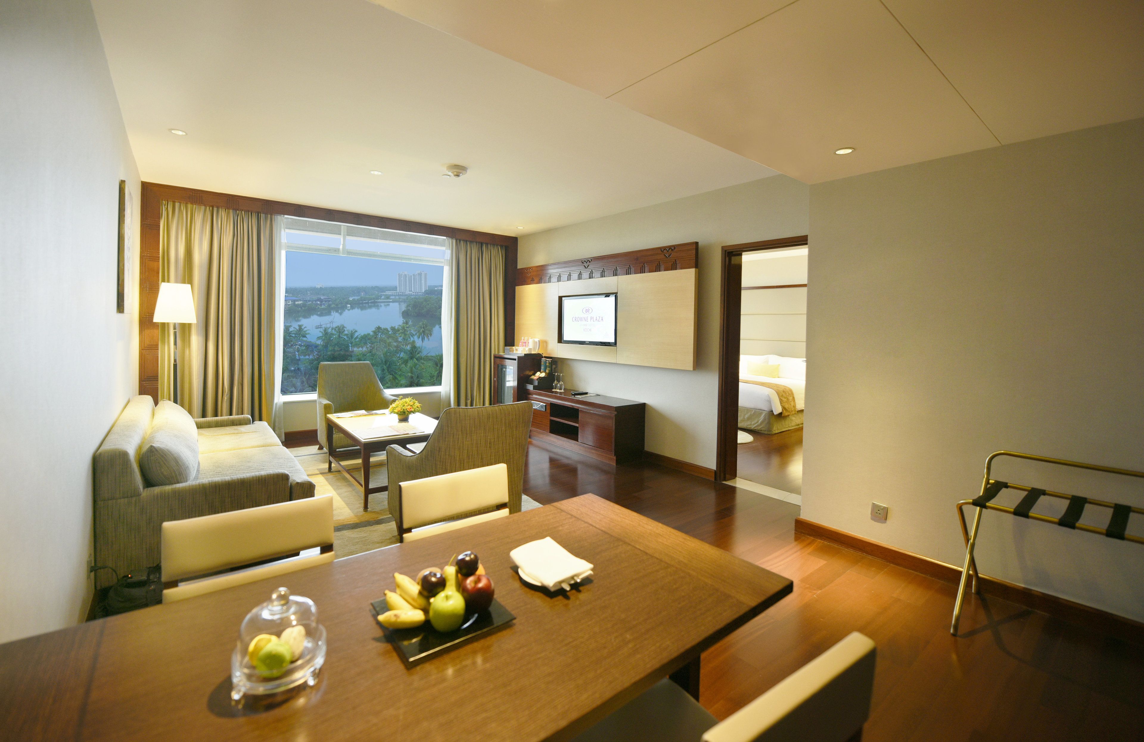 A hotel room with a dining table set with fruit, a sofa, a TV, and a view of the city.