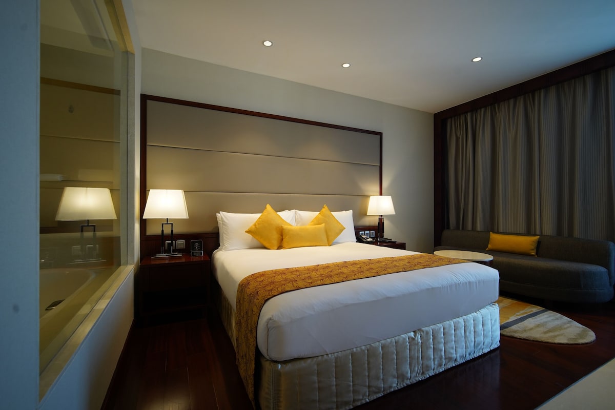 A hotel room with a large bed, two bedside lamps, a nightstand, a sofa, and a window with curtains.