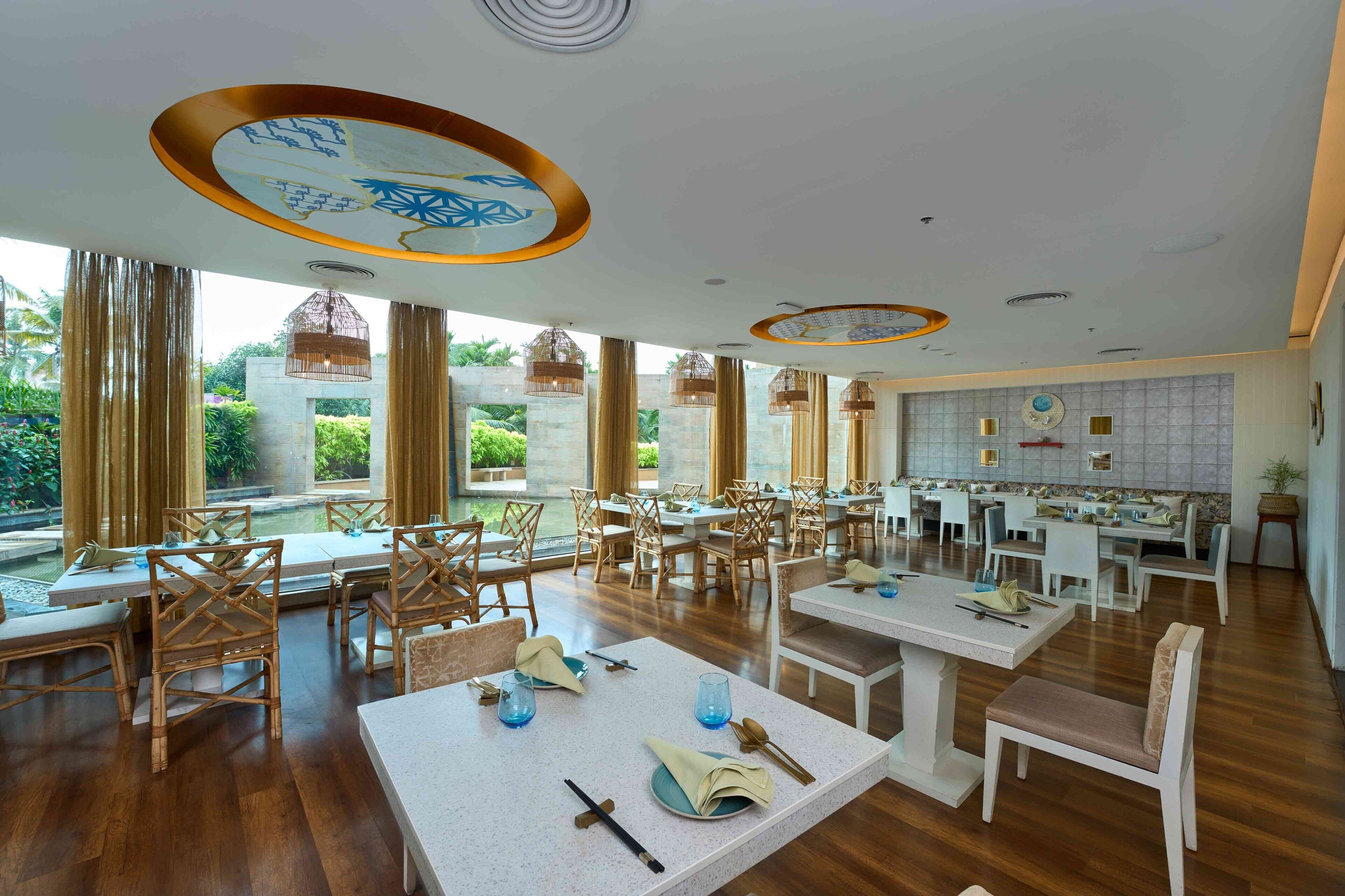A spacious restaurant with high ceilings, wooden flooring, and a view of greenery outside. Tables are set with white tablecloths and blue glassware.