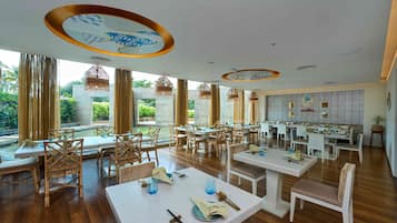 Un restaurante espacioso con techos altos, piso de madera y vistas al exterior con vegetación. Las mesas están dispuestas con manteles blancos y copas azules.