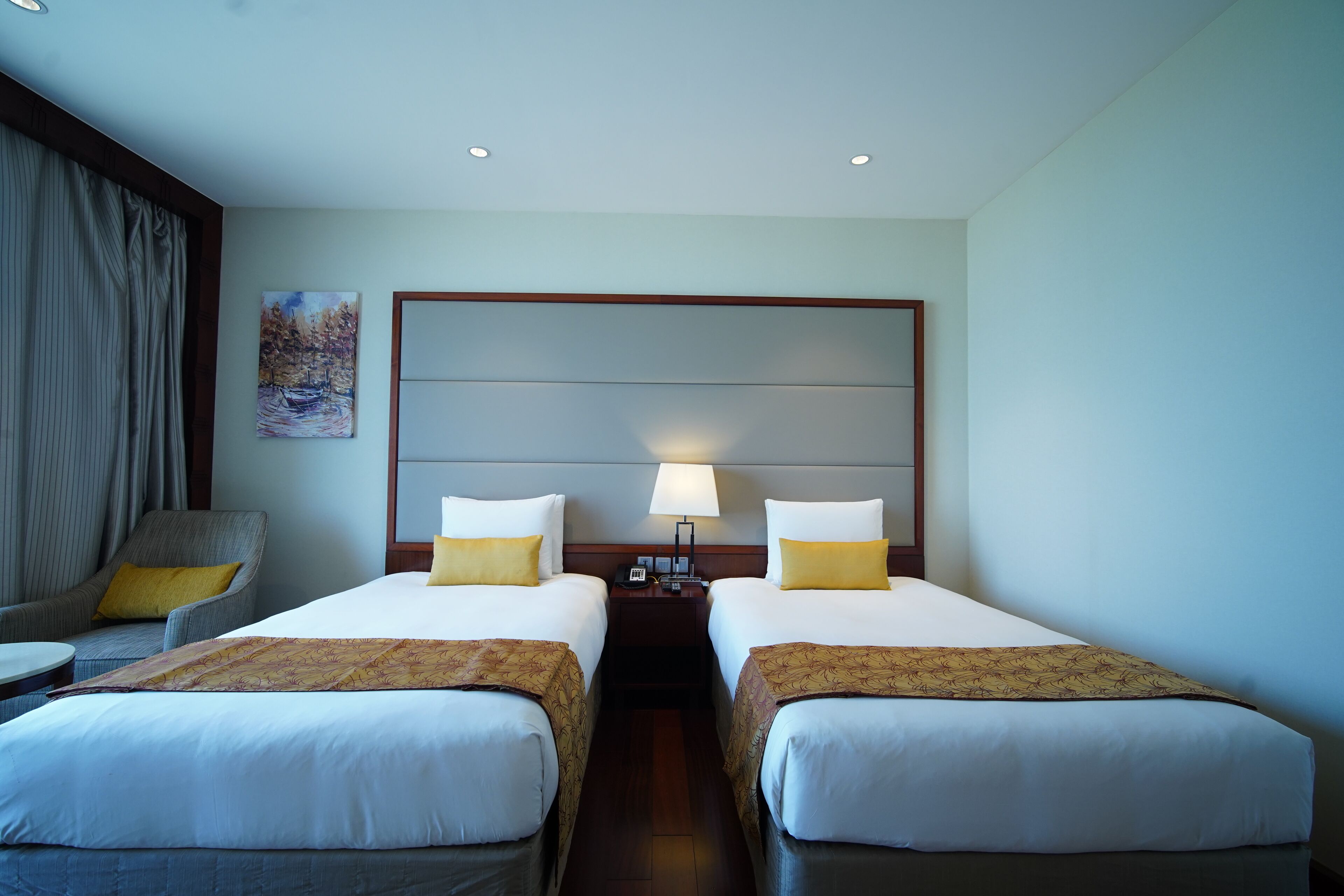 A hotel room with two beds, a nightstand with a lamp, a chair, and a painting on the wall.