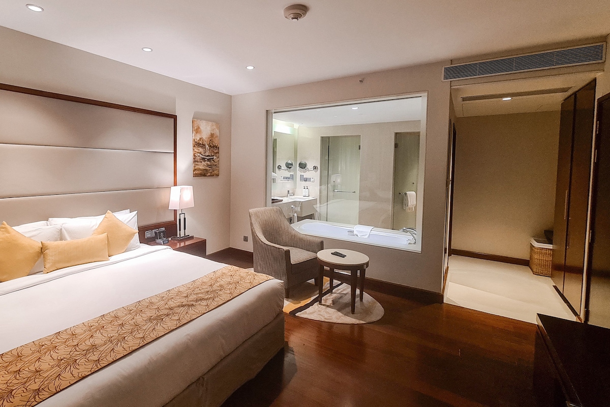 A hotel room with a large bed, a sitting area with a chair and table, a bathroom with a large mirror, and a door leading to another room.