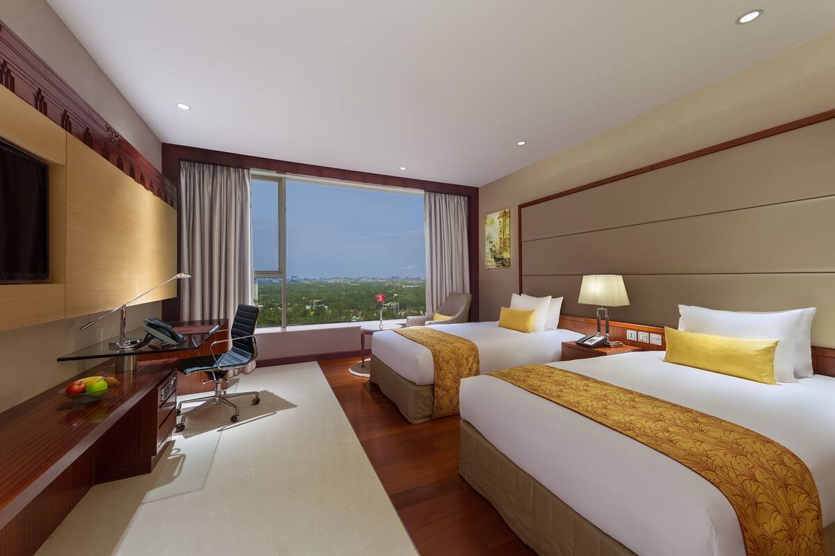 A hotel room with two beds, a desk, a chair, a TV, and a large window with a city view.