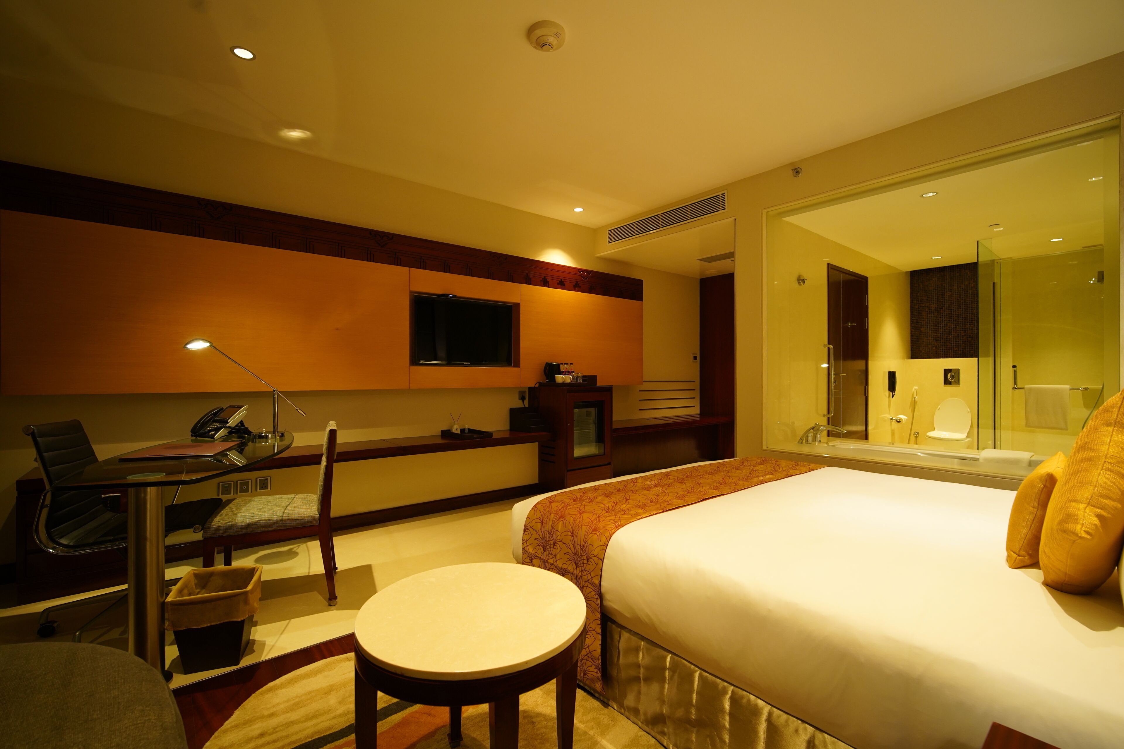 Habitación de hotel con cama, escritorio con silla, una mesita, televisión y baño visible a través de una puerta de cristal.