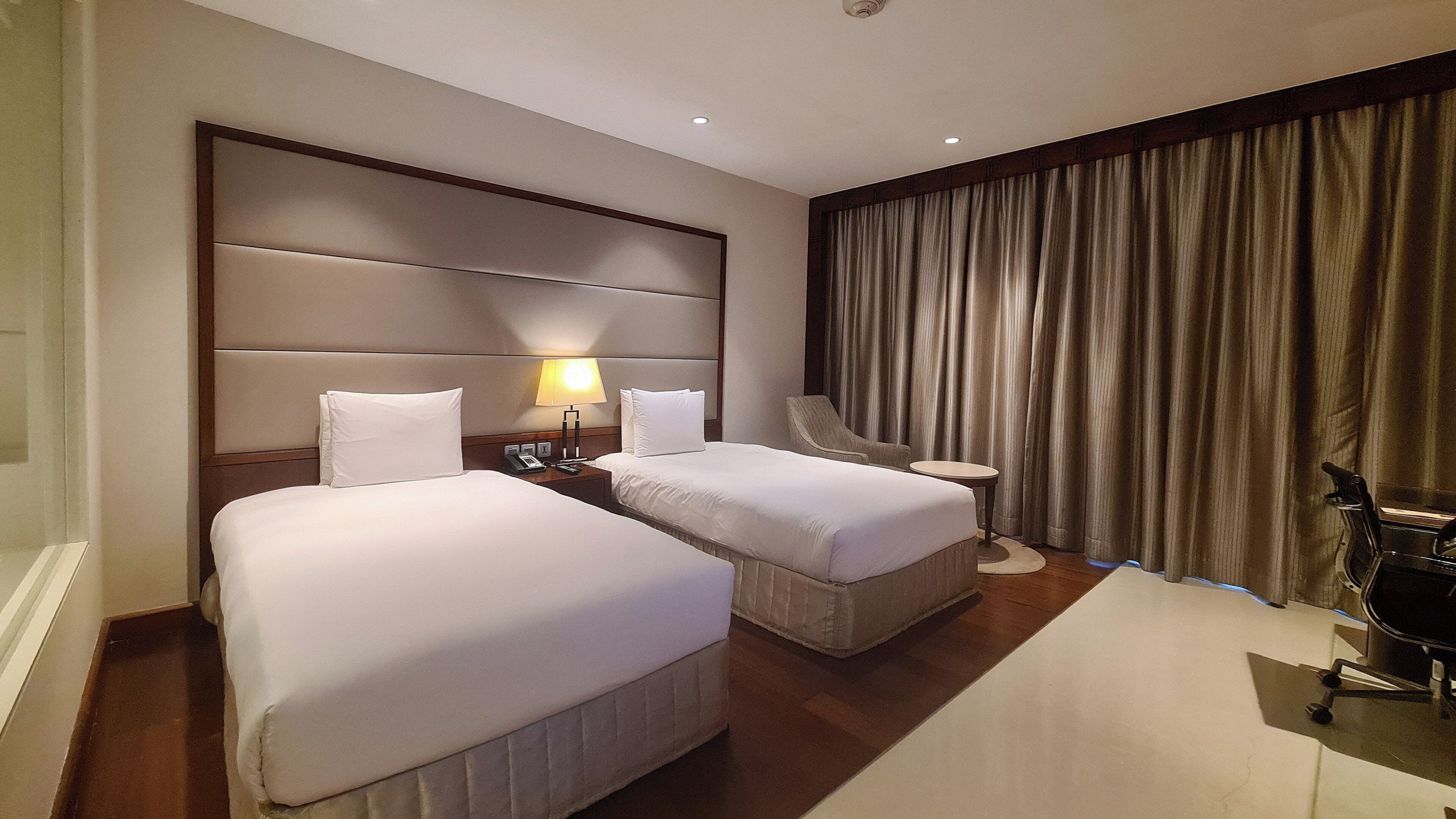 A hotel room with two beds, a nightstand, a chair, a small table, and a curtain.