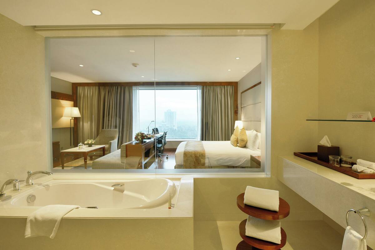 A hotel room with a large mirror, a bathtub, a desk with a chair, a bed, and a view of the city through the window.