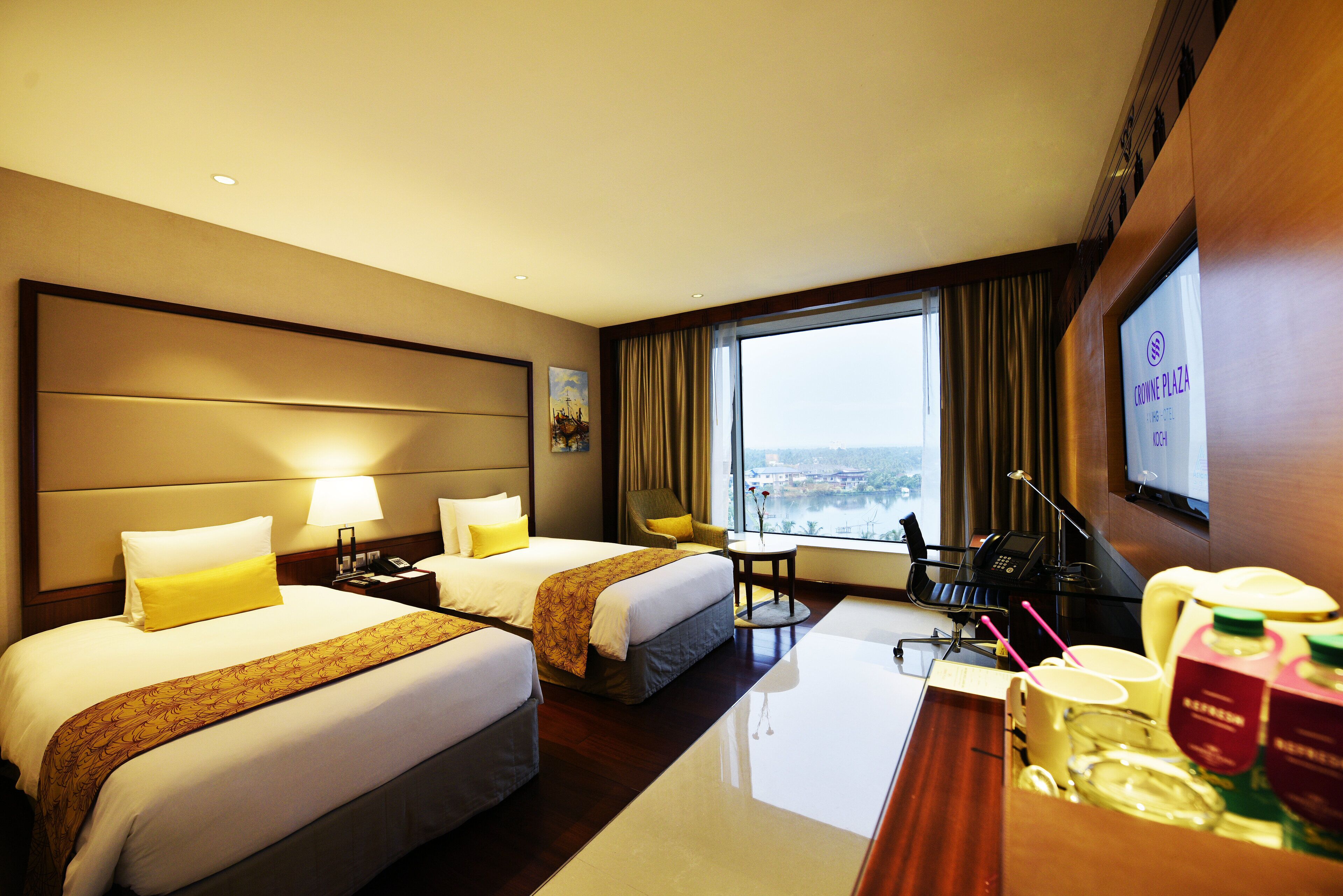 Habitación de hotel con dos camas, un escritorio con computadora, una silla y un ventanal con vista a la ciudad.