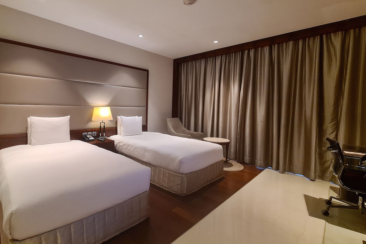 A hotel room with two beds, a nightstand with a lamp, a chair, a small table, and a large window with curtains.