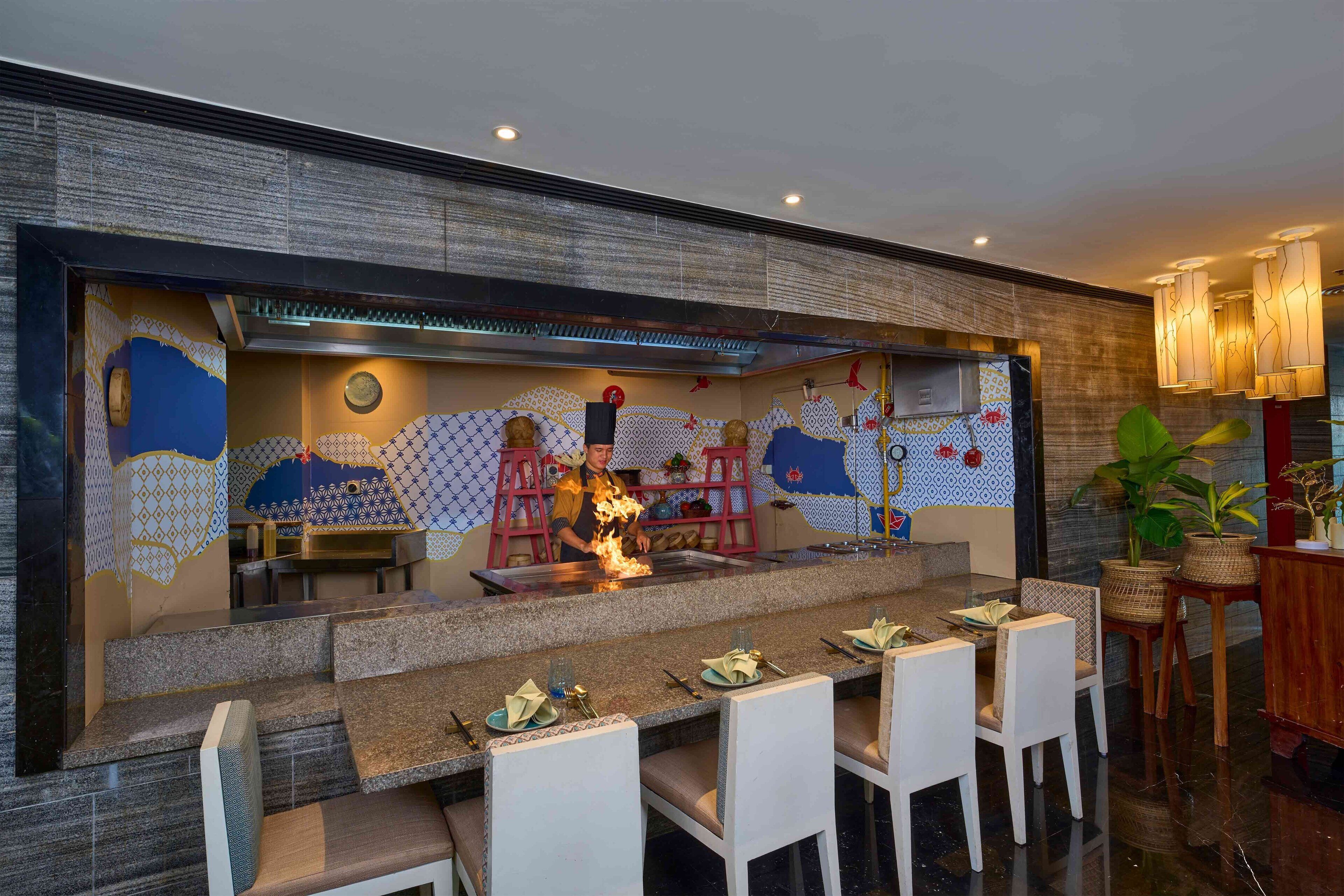 Un restaurant doté d’un espace de cuisine central, d’un comptoir de bar et de plusieurs tables à manger avec des chaises.