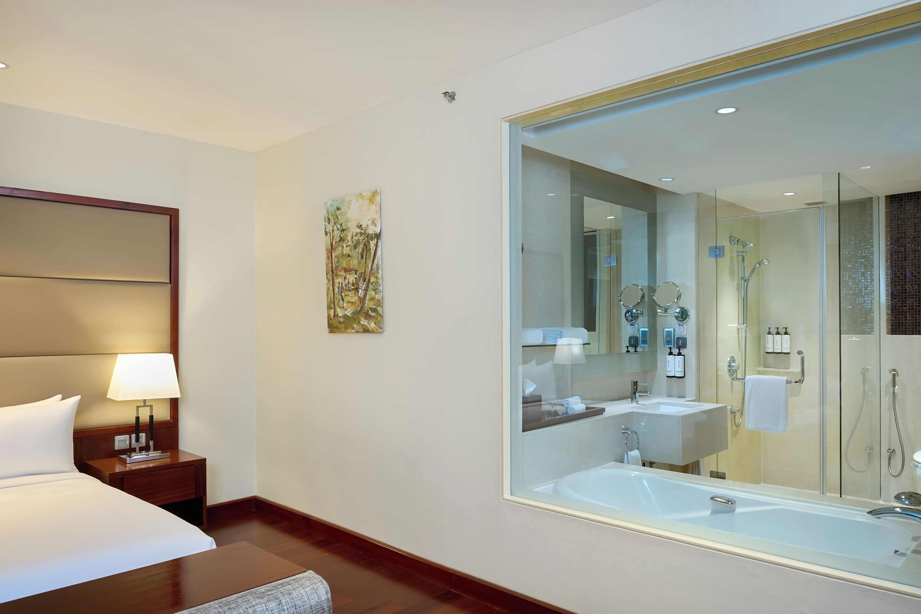 A hotel room with a large bed, a bedside table with a lamp, a painting on the wall, and a large mirror reflecting a bathroom with a bathtub and shower.